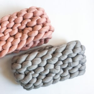 May include: Two chunky knit blankets, one in a light pink color and the other in a light gray color. The blankets are made of thick yarn and have a soft, fluffy texture.
