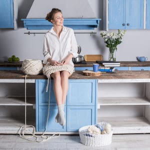 May include: A woman sits on a counter in a kitchen, knitting with white yarn. She is wearing a white shirt and a white knitted skirt. There are books and a cup on the counter behind her.