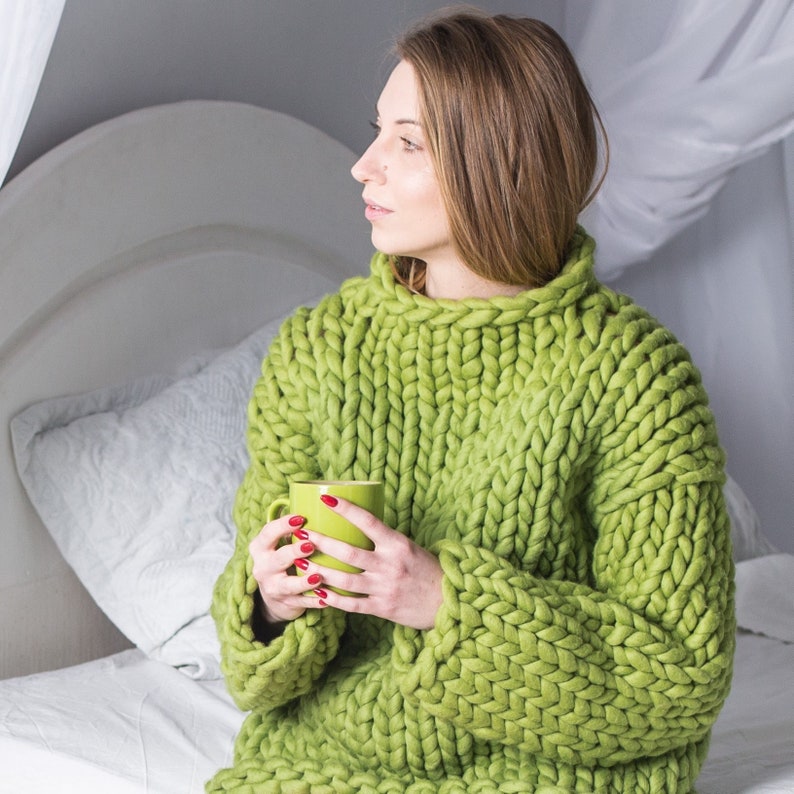 May include: A woman wearing a chunky knit green sweater, holding a green mug with both hands. She is sitting on a bed with white bedding.