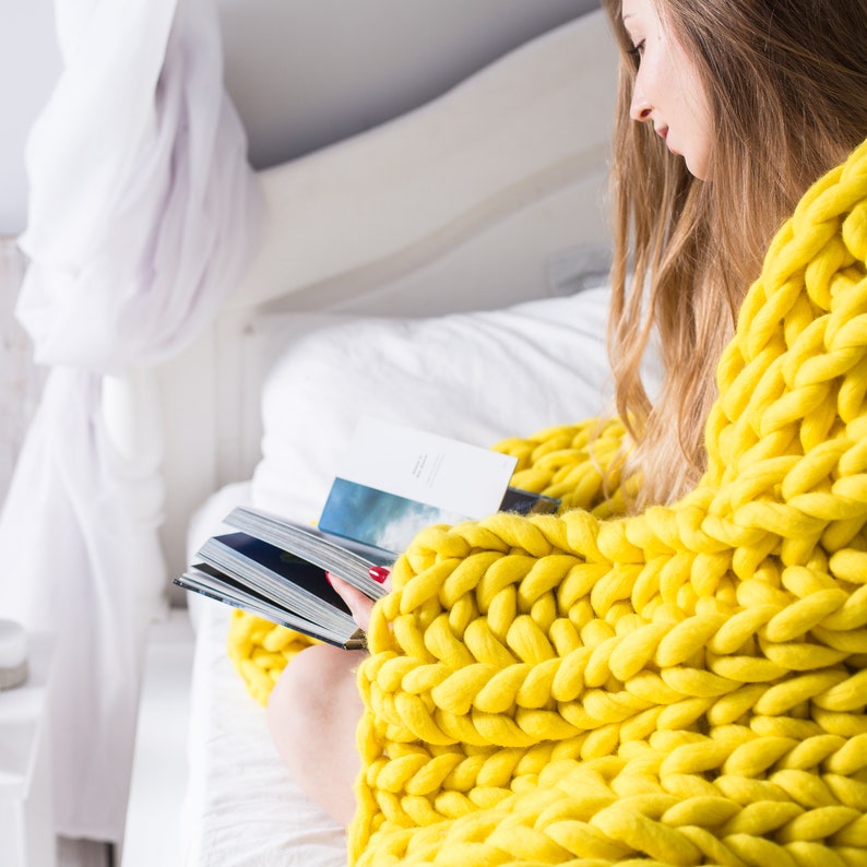May include: A woman relaxes on a bed, wrapped in a chunky knit yellow throw blanket, reading a book.