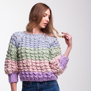 Woman wearing a pastel color block sweater hand knitted from chunky merino wool, with a cropped fit, round ribbed neckline and three-quarter raglan sleeves.