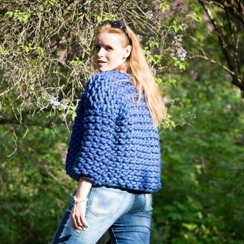 May include: A woman wearing a blue chunky knit cardigan with long sleeves and a relaxed fit. She is wearing blue jeans and has her hair pulled back.