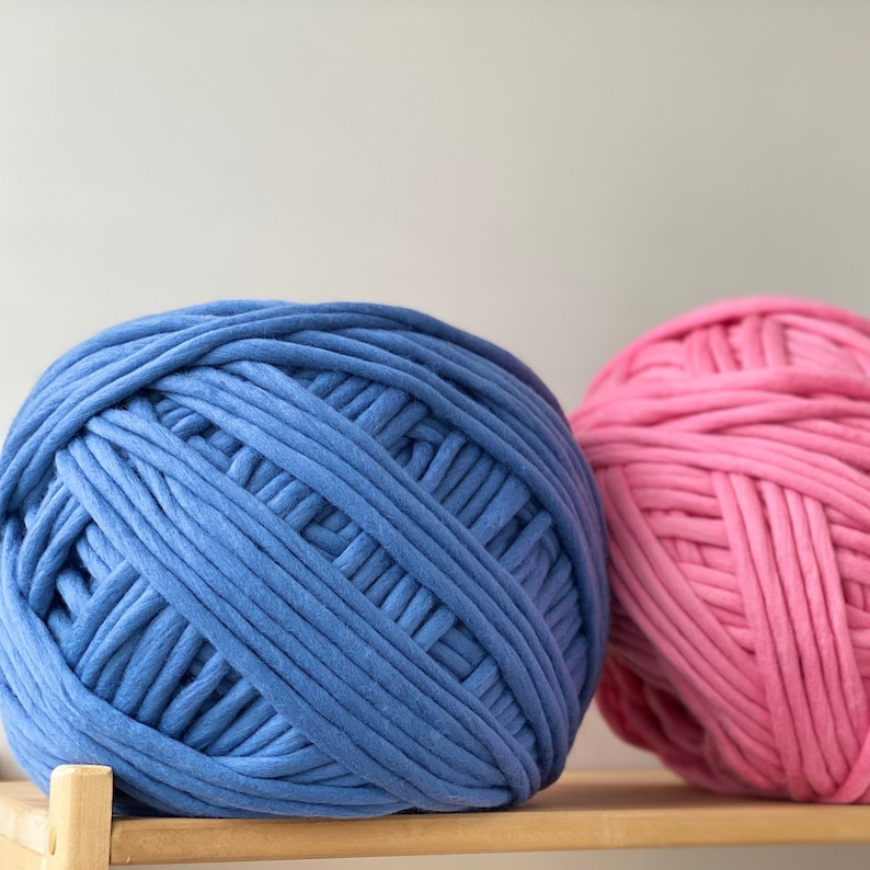 May include: Two balls of yarn, one blue and one pink, are sitting on a wooden shelf. The yarn is thick and fluffy, perfect for crocheting or knitting.