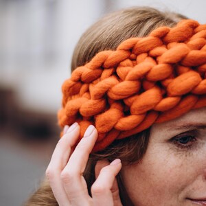May include: A close-up of a woman wearing a thick, orange knitted headband. The headband is made of chunky yarn and has a simple, textured design.