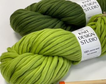 May include: Two skeins of thick, chunky yarn in shades of olive green and lime green. Each skein features a white paper label with the text KNIT DESIGN STUDIO, 100% merino wool, and 18.5 microns. Two wooden knitting needles are visible below.