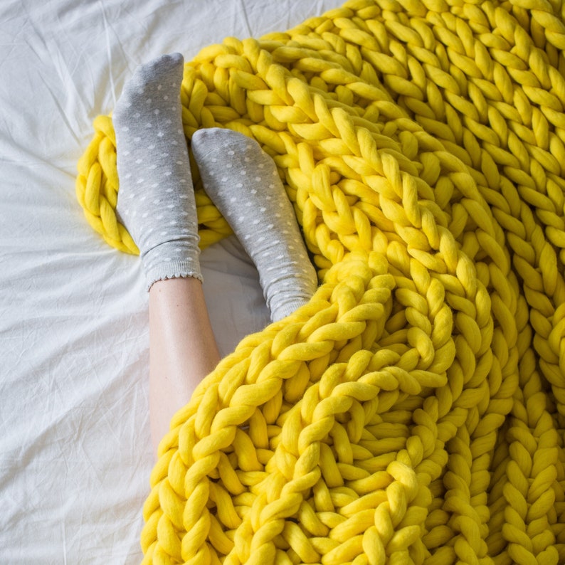May include: A yellow chunky knit blanket with a soft, fuzzy texture. The blanket is draped over a white bed and covers a pair of feet wearing grey socks.