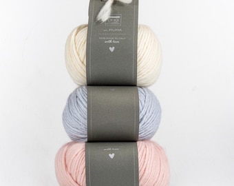 May include: A stack of three skeins of yarn in cream, light blue, and soft pink. Each skein is wrapped in a dark gray paper band featuring the text DHG, art. PIUMA, 100% MADE IN ITALY, with love, and a small heart icon. A white yarn tie sits on top.