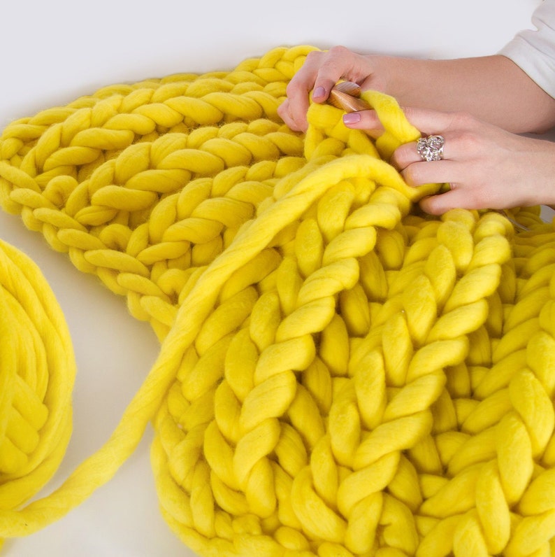 May include: A person is knitting a large, yellow, chunky knit blanket. The blanket is made with thick yarn and has a simple, textured stitch pattern. A ball of yarn is also visible in the image.
