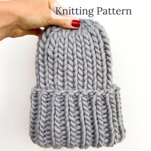 Op de afbeelding: Een dikke grijze gebreide muts met een omgeslagen rand. De hoed is gemaakt van dik garen en heeft een geribbelde textuur op de rand en een verticaal breipatroon op de kroon. De tekst "Knitting Pattern" staat in de linkerbovenhoek.