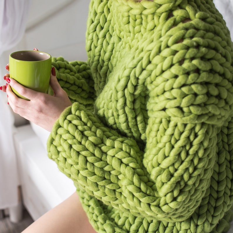 May include: A woman wearing a chunky knit green sweater. The sweater is made of thick yarn and has a loose, oversized fit.