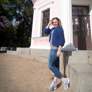 May include: A woman wearing a blue knitted cardigan, a striped shirt, blue jeans, and sneakers. She is standing in front of a pink building with a white trim.