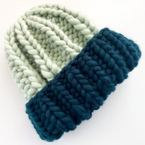 May include: A chunky knit beanie in two-tone colors. The top portion is a light green, while the folded brim is a deep teal. The hat is made of thick, soft yarn, perfect for cold weather. The knit pattern is a classic ribbed design.