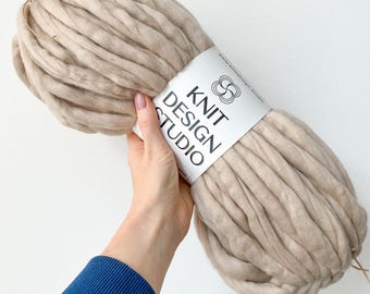 May include: A large skein of beige super chunky yarn. The yarn is held by a hand and has a white label with the text "KNIT DESIGN STUDIO" in black. The yarn is thick and appears soft, suitable for various knitting projects.
