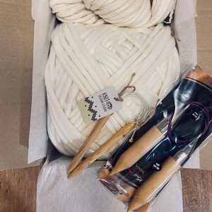 May include: A white chunky yarn ball with two wooden knitting needles and a set of three wooden knitting needles in a clear plastic package. The package has the text "Knit" and "80 cm" on it. The yarn ball is in a cardboard box with white tissue paper.