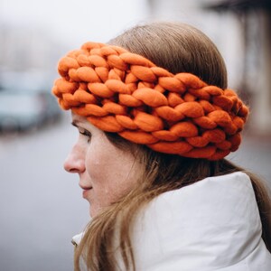May include: A woman wearing a bright orange knitted headband. The headband is thick and chunky, made with a large yarn.
