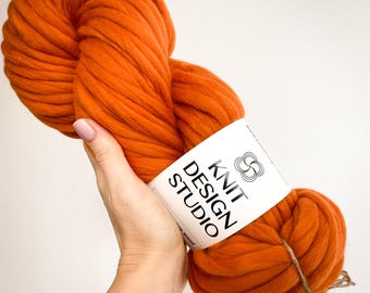 May include: A large skein of orange super chunky merino wool yarn. The yarn is held in a hand and has a white label that reads "KNIT DESIGN STUDIO". The yarn is tied with twine.