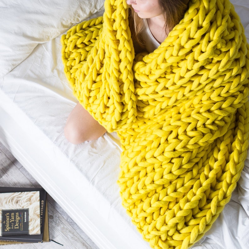 May include: A large, chunky knit throw blanket in a bright yellow color. The blanket is draped over a white bed and is made of thick, soft yarn.