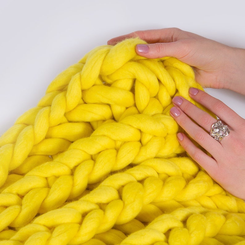 May include: A yellow chunky knit blanket made with thick yarn. The blanket is folded and a hand is resting on top of it.