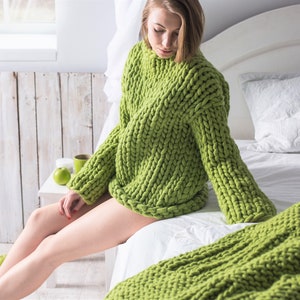 May include: A woman wearing a chunky knit green sweater sits on a bed with a green knitted throw blanket.