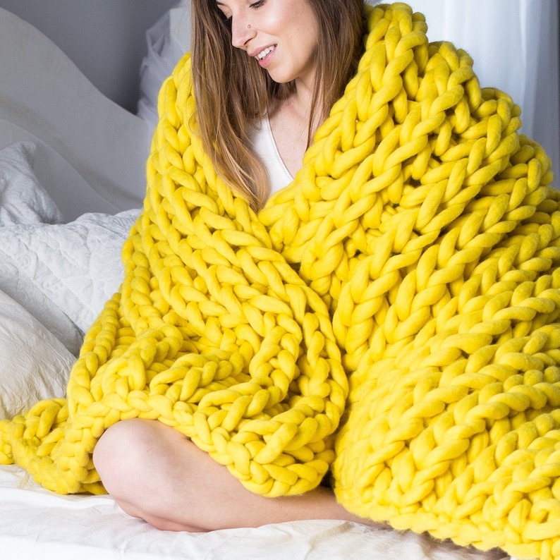May include: A yellow chunky knit blanket made of thick yarn. The blanket is draped over a person sitting on a bed.