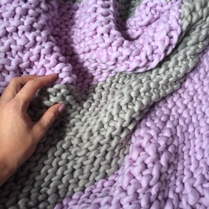 May include: A soft, chunky knit throw blanket in shades of lavender and gray. The blanket is made of thick yarn and has a textured, hand-knitted look.