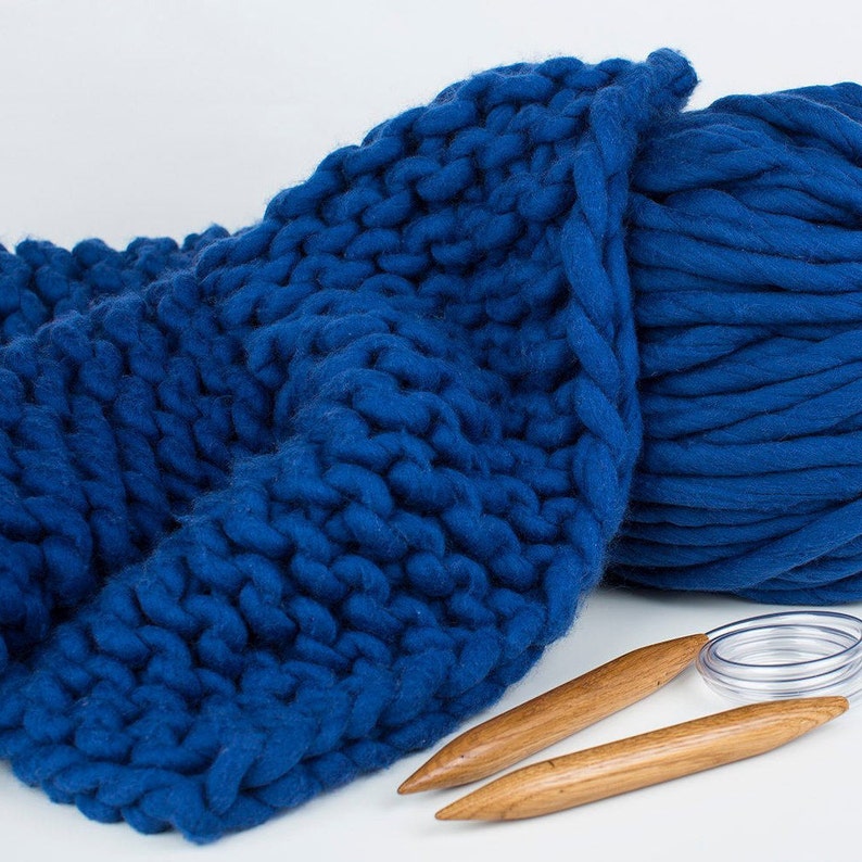 May include: A close-up of a blue knitted blanket made with chunky yarn. The blanket is folded in half and there are two wooden knitting needles next to it.