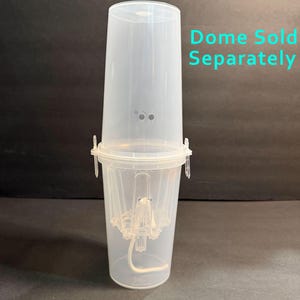 May include: A clear plastic cylindrical container with a locking lid and a central internal structure featuring a white cord. The text "Dome Sold Separately" is printed in light blue on the right side of the image, next to the upper section.