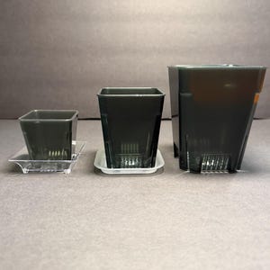 May include: Three black square plastic plant pots of varying sizes, each with a matching clear or white tray. The pots have drainage holes and are designed for indoor or outdoor use. The pots are arranged on a gray surface.