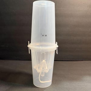 Puede incluir: Un recipiente de plástico transparente con forma cilíndrica, con tapa y base. El recipiente se compone de dos partes, con un mecanismo de cierre. La base contiene una cuerda blanca. El recipiente está diseñado para contener artículos.