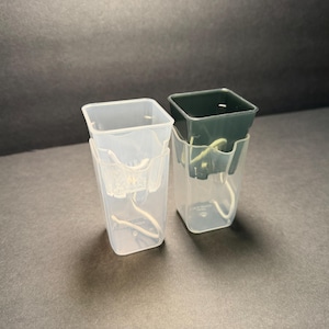May include: Two clear plastic plant pots with a square top and a clear plastic holder. One pot is empty, and the other has a dark green pot inside. The pots are on a dark gray surface.
