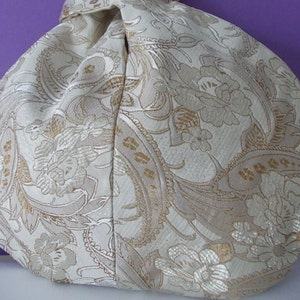May include: A cream-colored, paisley-patterned bag with gold accents. The fabric has a subtle sheen, and the bag is gathered at the top. The floral and paisley design is visible across the surface of the bag.