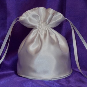 May include: A white satin drawstring bag with white ribbon ties. The bag is on a purple background.