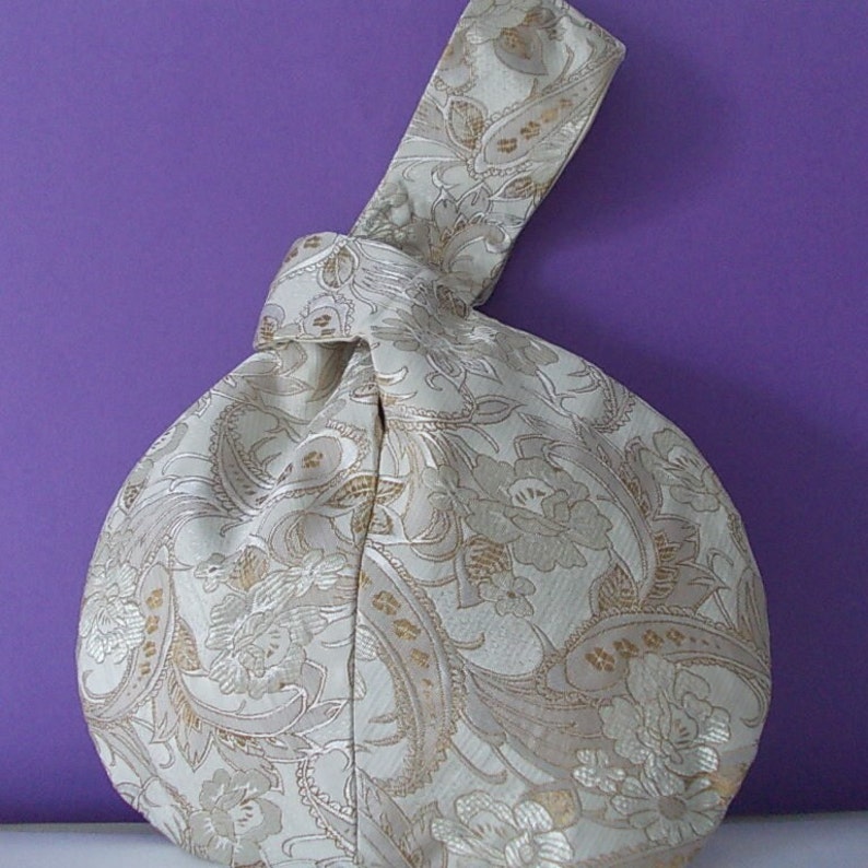 May include: A cream-colored, paisley-patterned handbag with a knotted handle. The bag features a repeating floral and scroll design in gold and cream tones. The background is a solid purple.