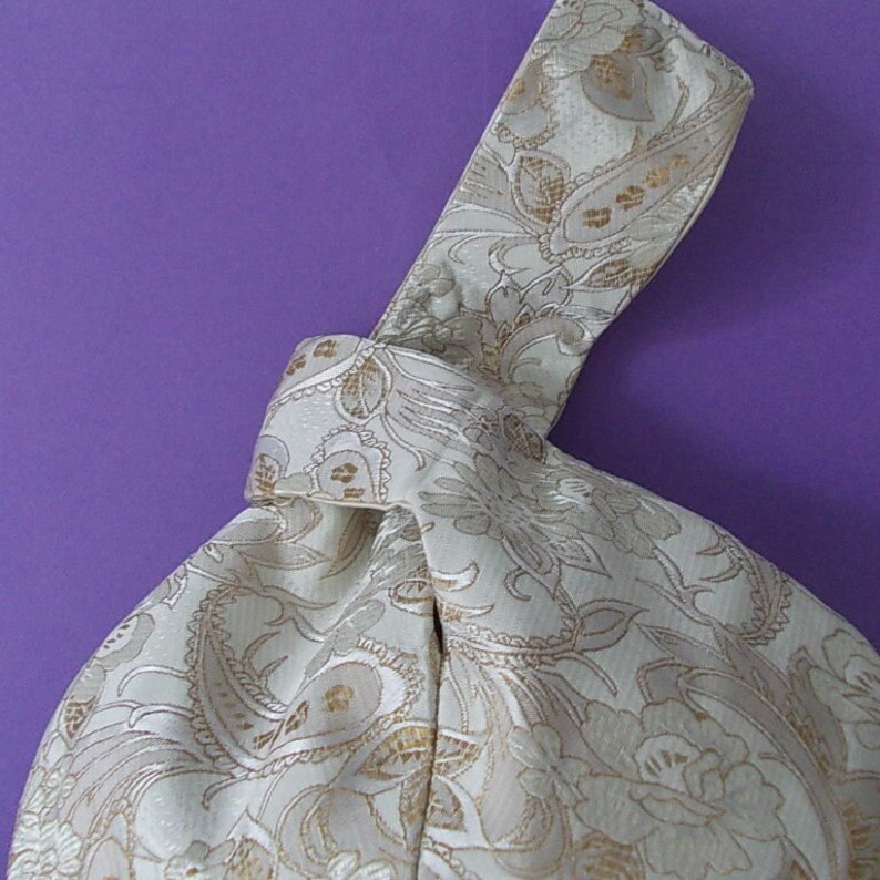 May include: Ivory-colored fabric bag with a knotted handle. The bag features a repeating floral and paisley pattern in gold and cream tones. The background is a solid purple.