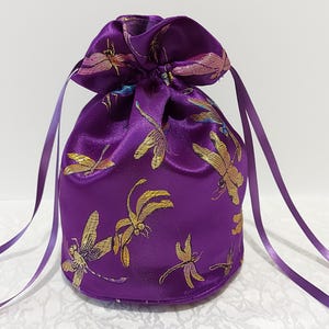 May include: A purple satin drawstring bag with a gold dragonfly pattern. The bag has a long purple ribbon tie.