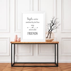 Könnte beinhalten: Ein weißer gerahmter Druck mit einem schwarzen Zitat, das lautet "Engel existieren, aber manchmal haben sie keine Flügel und werden FREUNDE genannt". Der Druck liegt auf einem Holztisch mit einer Vase mit Zweigen und zwei rosa Dekorationsgegenständen.