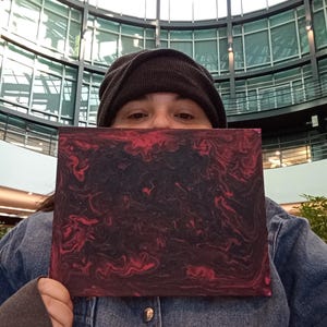 Può includere: Una tela rettangolare con un design astratto a spirale nei toni del rosso e del nero. L'opera d'arte è tenuta davanti a una persona che indossa un berretto scuro e una giacca di jeans, su uno sfondo architettonico moderno.