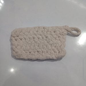 May include: An off-white, rectangular crocheted washcloth with a hanging loop. The washcloth has a textured, nubby surface, made from a soft, thick yarn. The loop is made of the same material.