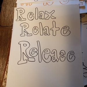 May include: A white paper with the words "Relax, Relate, Release" in large, black, bubble letters. The words are stacked vertically. Other papers with handwritten words are visible in the background.