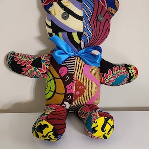 May include: A colorful patchwork teddy bear with a blue bow. The bear is made from different fabrics with various patterns, including floral, geometric, and abstract designs. The bear has black button eyes.