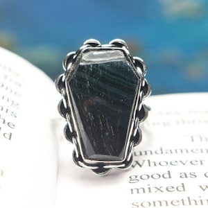 May include: A silver ring with a black gemstone in the shape of a coffin. The ring is on a white surface.