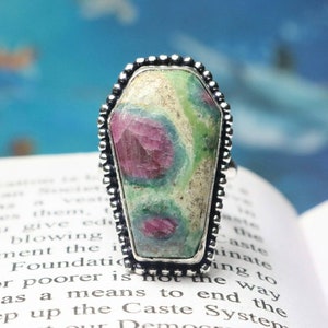 Peut inclure: Une bague en argent avec une pierre précieuse verte et rose sertie dans un cadre en forme de cercueil. La pierre précieuse présente un motif tourbillonnant de vert, de rose et de blanc.