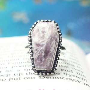 Pode incluir: Um anel de prata com uma configuração em forma de caixão com uma gema púrpura e branca.