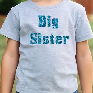 以下が含まれることがあります： 薄いグレーのTシャツに、青いグリッター文字で「Big Sister」と書かれています。