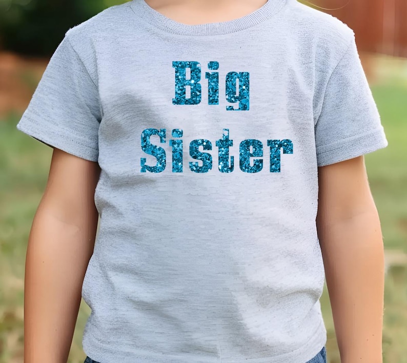 以下が含まれることがあります： 薄いグレーのTシャツに、青いグリッター文字で「Big Sister」と書かれています。