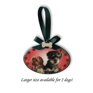 May include: Oval-shaped Christmas ornament with a red background and paw print design. Two dogs are pictured on the ornament, with the text "Pipsqueak & Mozi" below them. The ornament has a green ribbon and a bone-shaped charm.