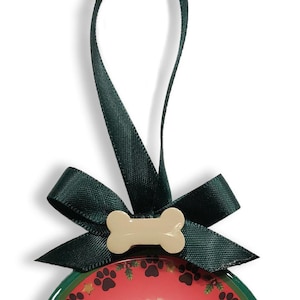 May include: A green and red Christmas ornament with a bone-shaped hanger and a green ribbon bow. The ornament features paw prints and holly.