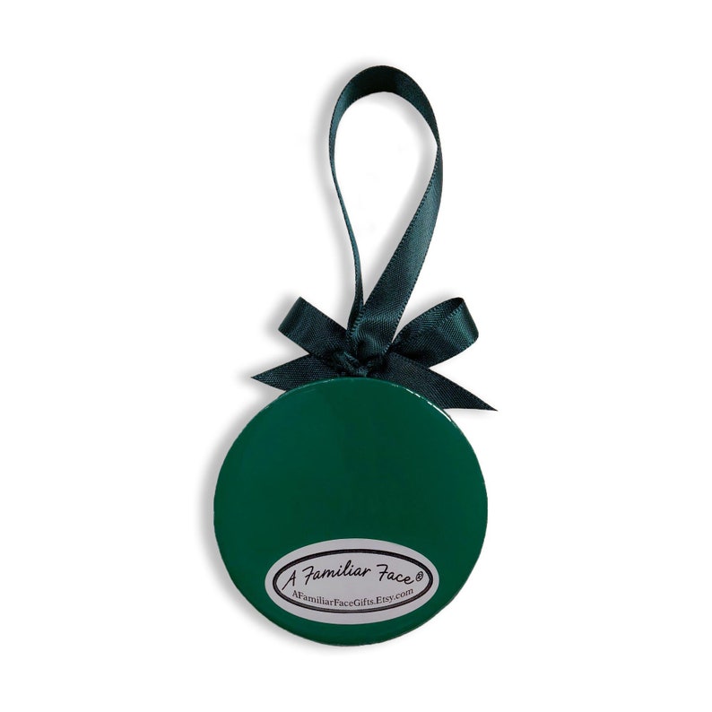May include: A green round ornament with a black ribbon and bow. The ornament has a white label with the text "A Familiar Face&reg;" and "AFamiliarFaceGifts.Etsy.com".