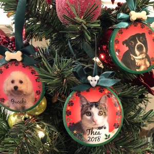 May include: Three personalized Christmas ornaments featuring pet portraits. A white dog named "Gucci", a black and white dog named "Xena", and a tabby cat named "Thea 2018". Each ornament has a red background with paw prints and a green border.