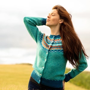 Women standing in a field of wheat wearing an Aqua colour wool fair isle cardigan. Crathie pattern with orange and teal.